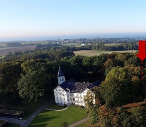Luchtbeeld van een pand met een groot wit gebouw, omgeven door bomen en velden. Een rode pijl wijst naar een kleiner gebouw, aangeduid als een sauna.