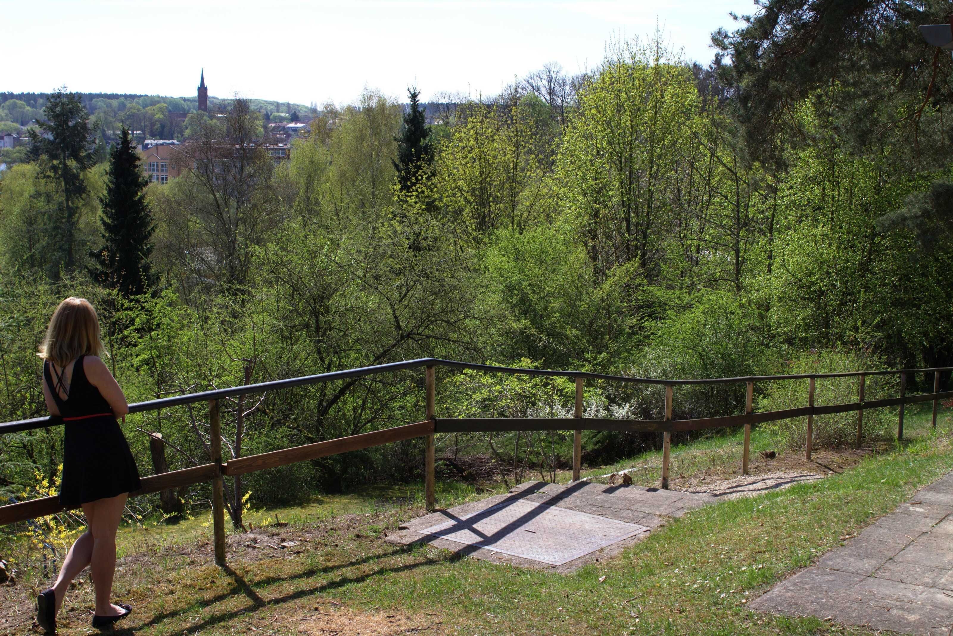 A woman walking on a wooden pathway with a metal railing, surrounded by greenery and a distant building.
