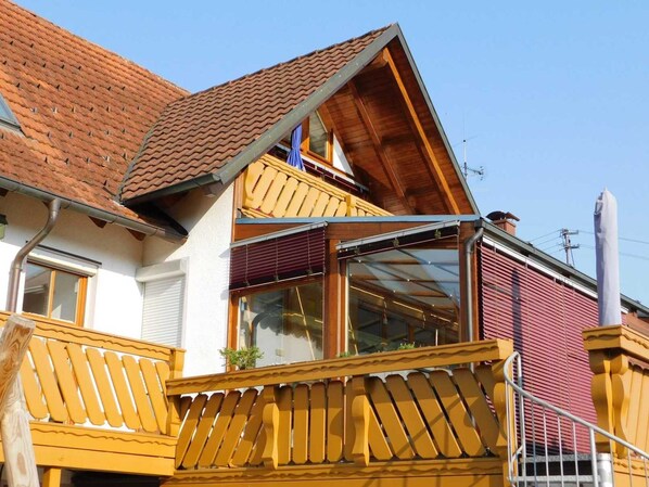 A two-story house with a red-tiled roof, a balcony with yellow railings, and a large window.