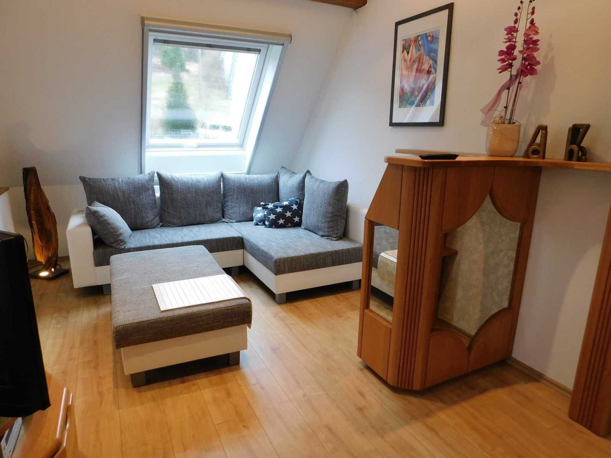 Ein modernes Wohnzimmer mit einem Ecksofa, einem Holztisch, einem kleinen Beistelltisch, einem gerahmten Bild an der Wand und einem Fenster mit Aussicht.