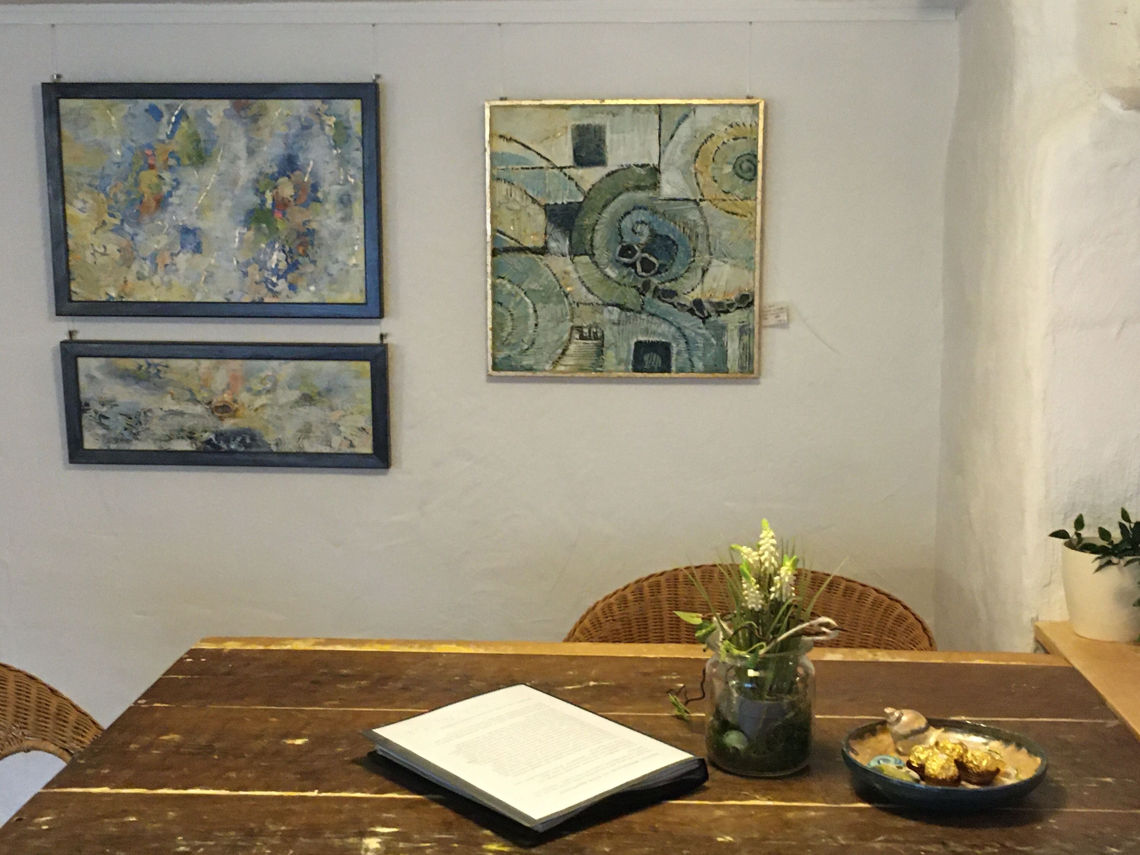 Un comedor con una mesa de madera, dos cuadros abstractos enmarcados, un florero con flores y un bol de bombones.