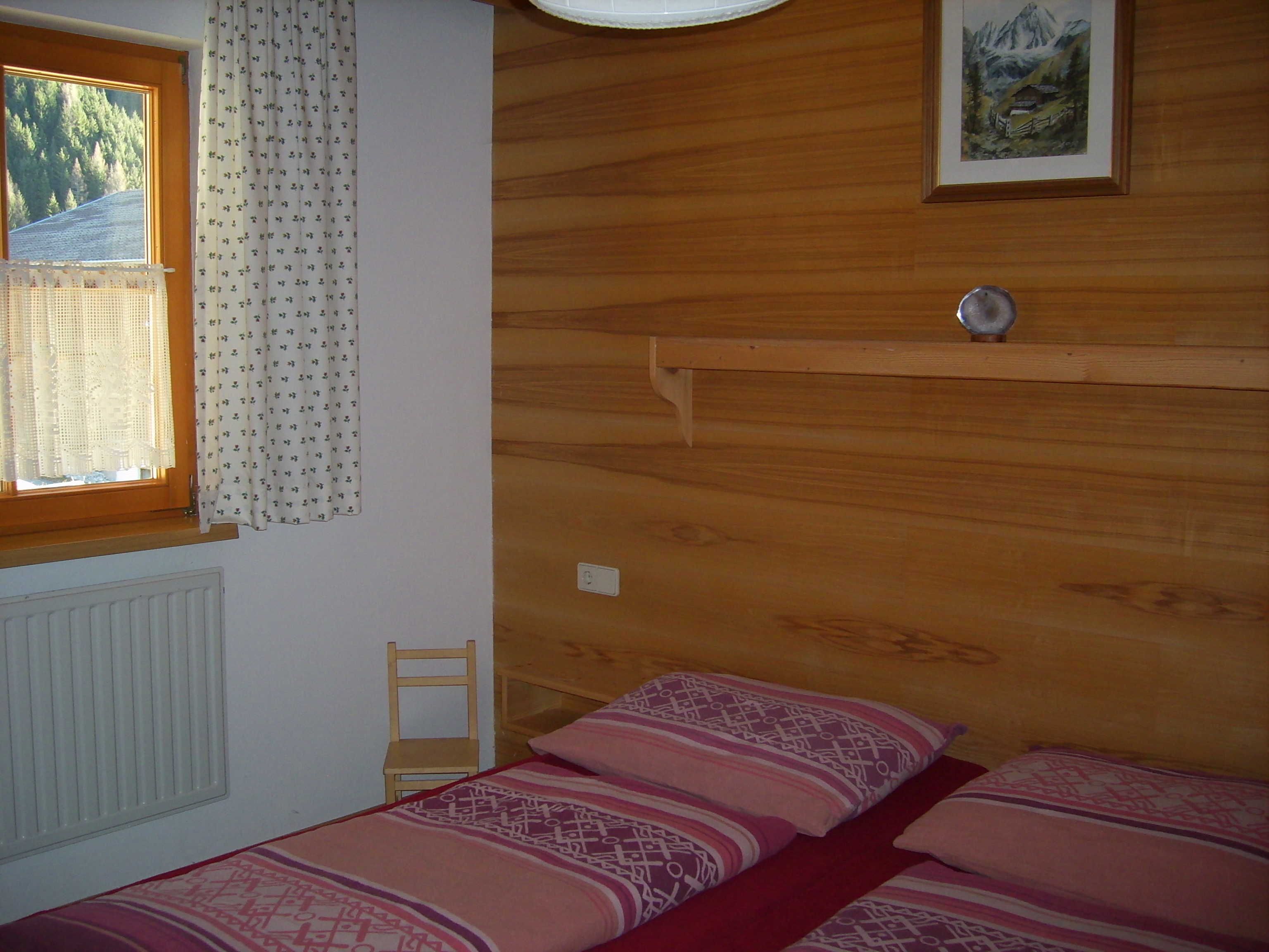 A bedroom with a wooden headboard, a bed with patterned bedding, a chair, a window with curtains, and a painting on the wall.