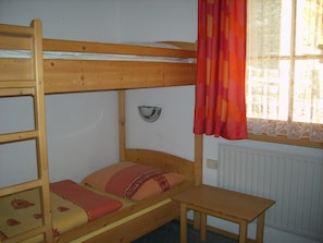 Una cama litera con una mesita pequeña, una ventana con cortinas y un radiador.