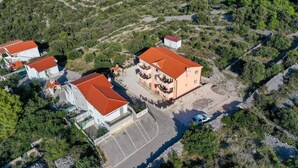 Vista aérea de una zona residencial con varias casas, incluyendo un edificio de dos pisos con techo rojo y balcones, rodeado de vegetación y un área de estacionamiento.