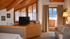 Ein gemütliches Zimmer mit Holzboden, einem Fernseher auf einer Holzkommode, einem Sofa und einem Fenster mit Vorhängen.