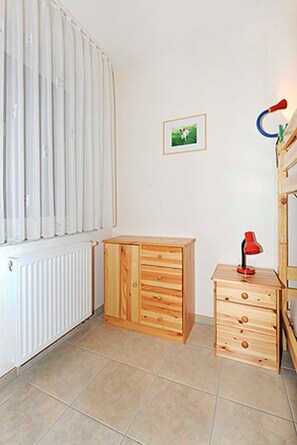 Quarto com radiador, um armário de madeira com gavetas, uma pequena escrivaninha com abajur vermelho e um quadro na parede.