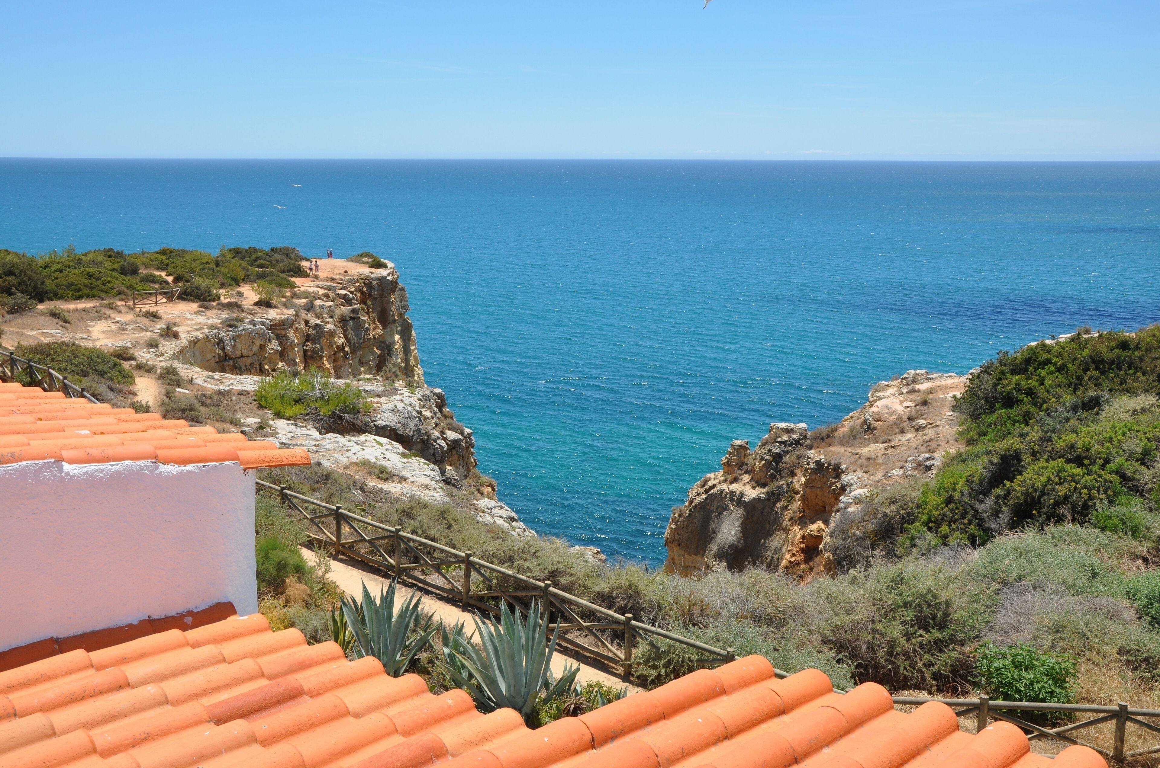 A coastal view with terracotta-roofed buildings, rocky cliffs, and the sea.