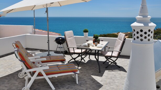 Um terraço com cadeiras de lounge brancas, uma mesa posta para refeições e vista para o mar.
