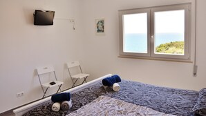 Quarto com cama, duas cadeiras, uma mesinha, TV na parede e janela com vista para o mar.