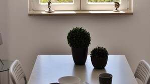 Mesa de jantar com pratos, tigelas e copos, uma planta em vaso e vista para o mar através de uma janela.