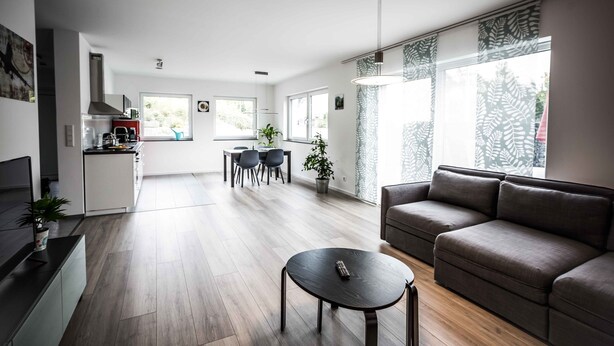 Un salon moderne avec un canapé, un coin repas avec des chaises, un espace cuisine et de grandes fenêtres avec des rideaux.