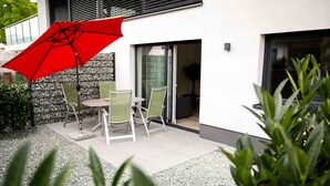 Une terrasse extérieure avec une table et des chaises, un parasol rouge et un bâtiment moderne doté d’une porte en verre.