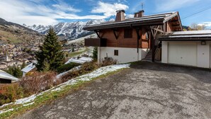 Una cabana de muntanya amb façana de fusta, un garatge blanc i una vista d'una vila i muntanyes nevades al fons.