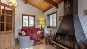 Un saló d'estar acollidor amb una llar de foc, bigues de sostre de fusta, un sofà vermell i un butac armchair gris.