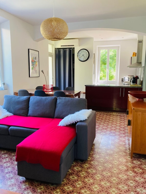 A living room with a grey sofa, a red throw blanket, and a woven pendant light. There is a dining area with a table and chairs, and a kitchen with a microwave and cabinets.