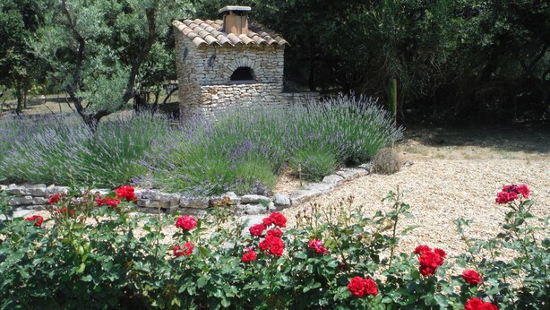 A stone building with a tiled roof, surrounded by a garden with red roses and lavender plants.