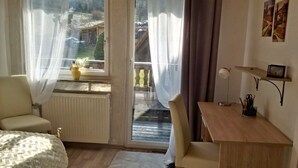Quarto com cama, escrivaninha, cadeira, radiador, janela com cortinas e vista para um deck de madeira lá fora.