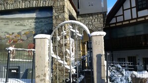 Um portão coberto de neve com o número '39' em uma parede de pedra, um portão de metal com desenhos elaborados e um edifício com fachada de madeira e estuque em segundo plano.