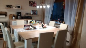 Uma sala de jantar com uma mesa de madeira, cadeiras brancas e um candelabro. Aparelhos de cozinha podem ser vistos ao fundo.