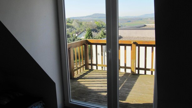 Blick vom Balkon mit Holzwertung auf eine Landschaft mit Hügeln und blauem Himmel.