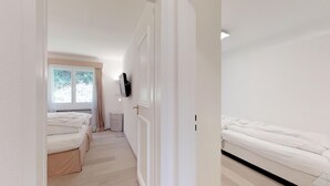 Un pasillo estrecho que conduce a dos camas individuales, una ventana y un televisor en la pared.