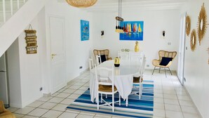 Comedor con una mesa blanca, sillas y un tapete azul a rayas. Se ve una escalera con barandillas blancas y una puerta con un cartel.