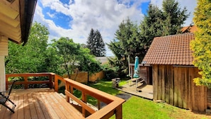 Une terrasse en bois avec un banc, un petit abri de jardin et une cour gazonnée avec des arbres.