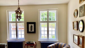 A room with two large windows, a radiator, a patterned armchair, a small table with books, and a chandelier. - Small castle Lanke (Lanke)