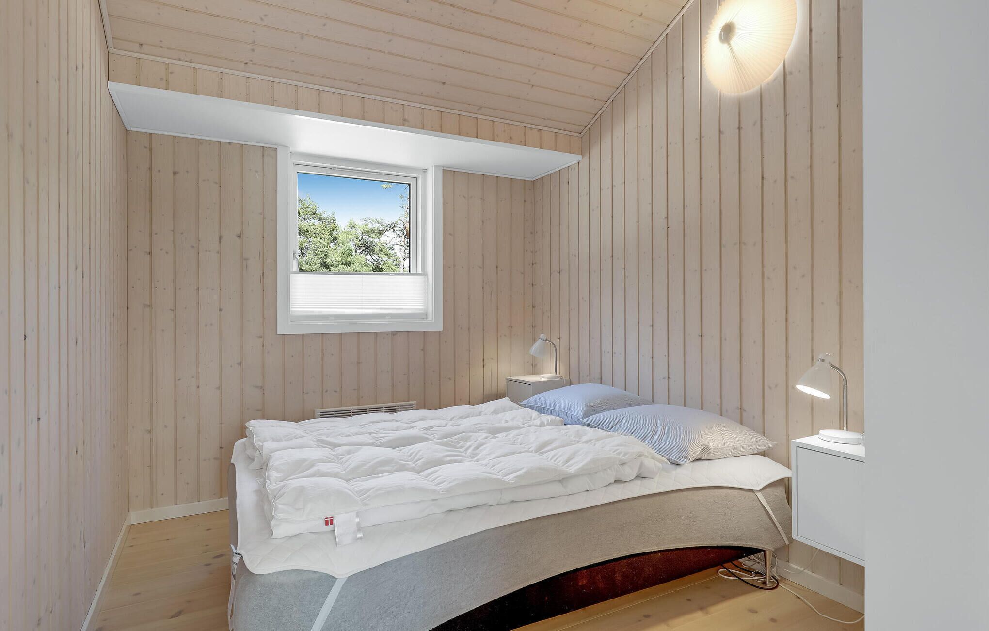 A bedroom with a sloped ceiling, a window with a view of greenery, a bed with white bedding, and a bedside table with a lamp.