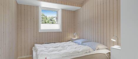 A bedroom with a sloped ceiling, a window with a view of greenery, a bed with white bedding, and a bedside table with a lamp.