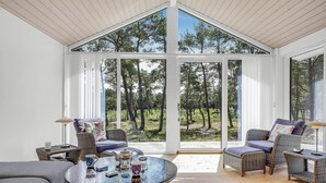 Une pièce spacieuse avec un plafond haut et de grandes fenêtres donnant sur une forêt. On y trouve des fauteuils en osier, une table ronde avec un service à thé et un canapé.