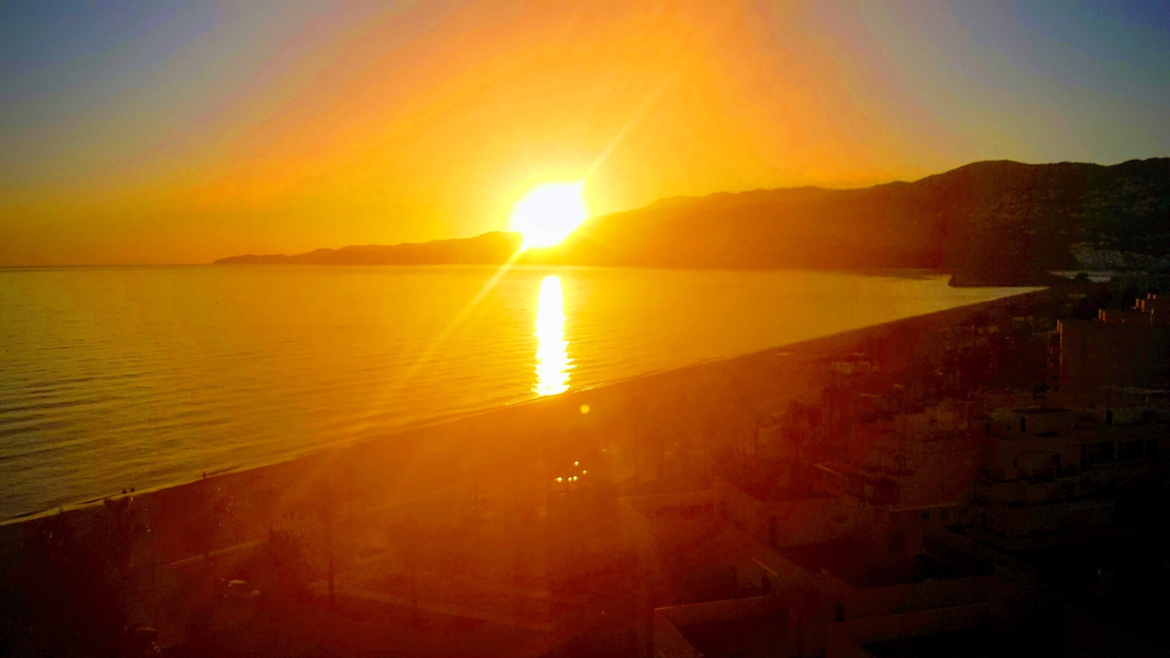 Un atardecer sobre un mar tranquilo con una playa y edificios al frente.