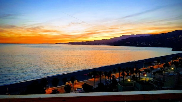 Vista costera con un atardecer sobre el agua, un paseo marítimo con palmeras y montañas a la distancia.