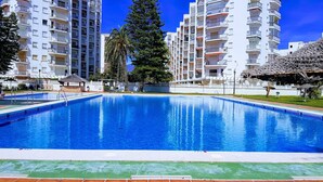 Una piscina rodeada de edificios residenciales altos, una palmera y una estructura con techo de paja.