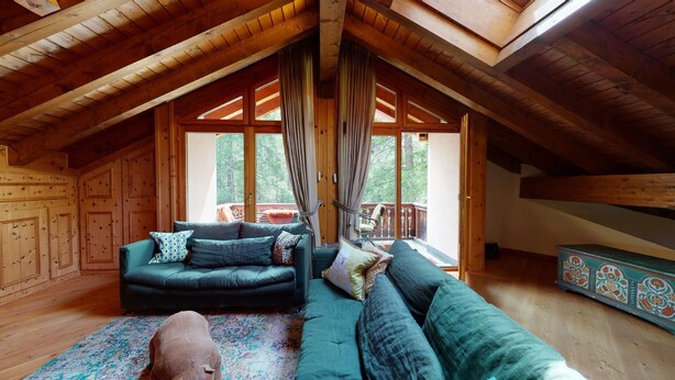 A cozy attic room with a wooden ceiling, a green sofa set, a patterned rug, and a view of the outdoors through large windows.
