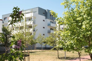 A modern apartment building with balconies, surrounded by young trees and a grassy area.