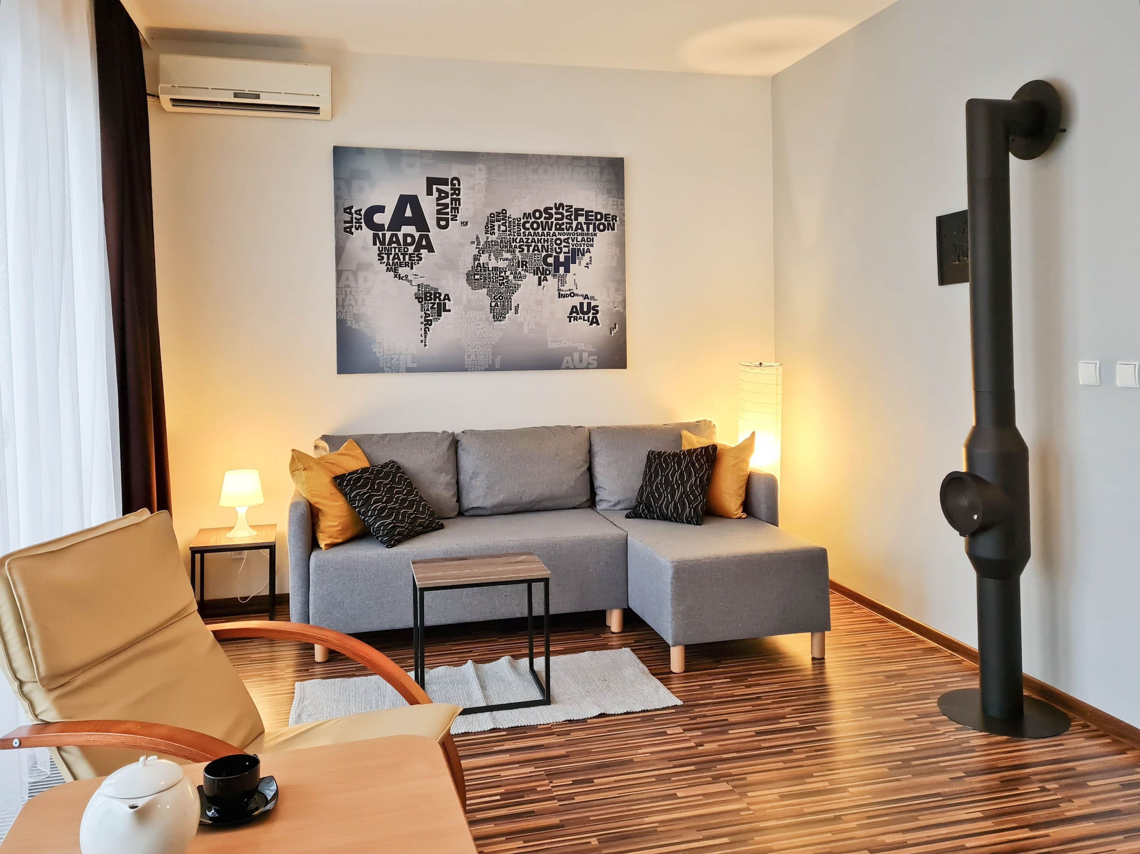 Ein modernes Wohnzimmer mit einem grauen Sofa, zwei Beistelltischen mit Lampen, einem Holztisch als Couchtisch und einem beigen Sessel, sowie einer Weltkarte als Wandbild.