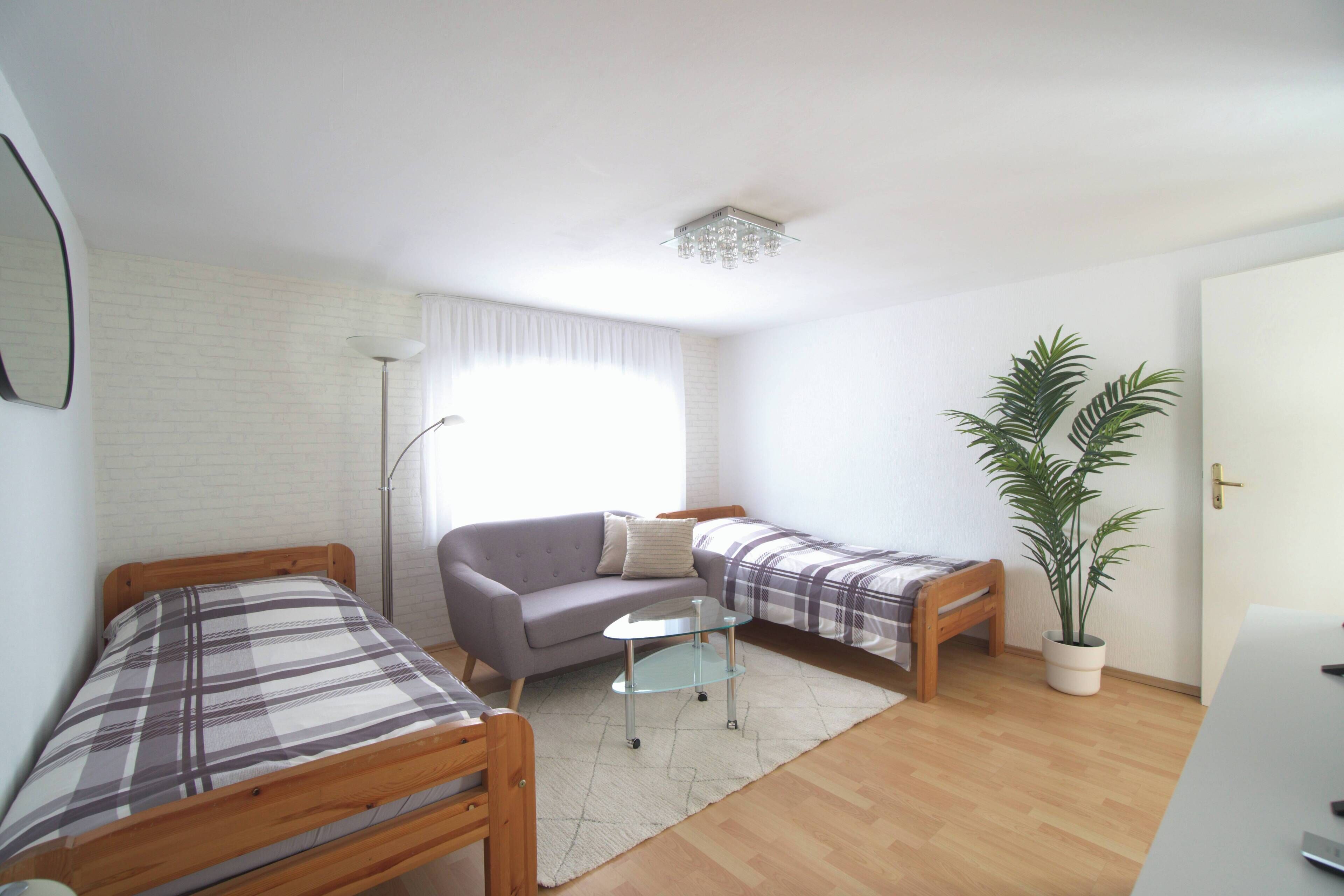 A small, well-lit room with two single beds, a sofa, a small table, a potted plant, and a door.