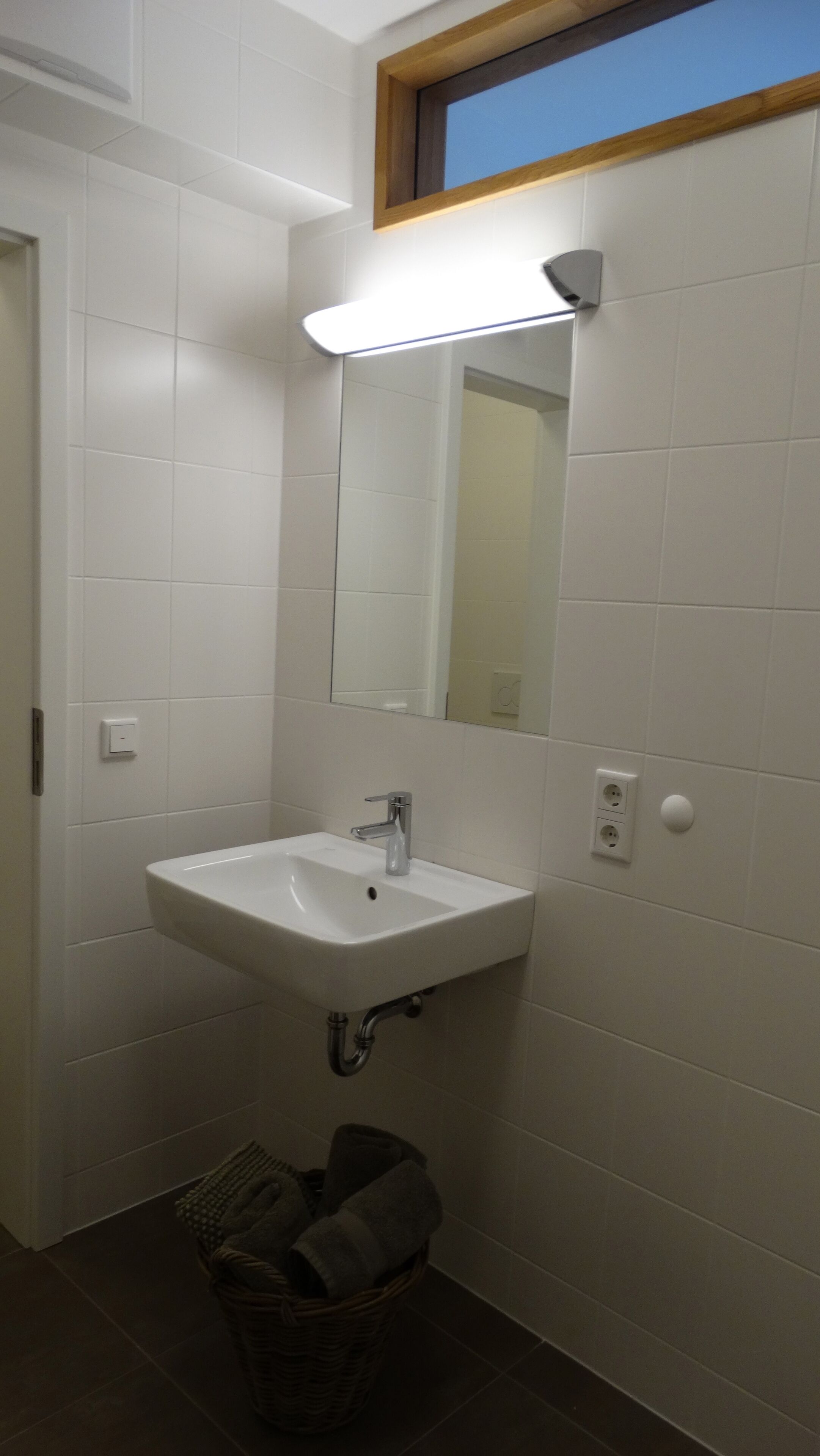 Un baño con un lavamanos blanco, un espejo y una lámpara sobre el espejo. Hay una cesta con toallas en el suelo.