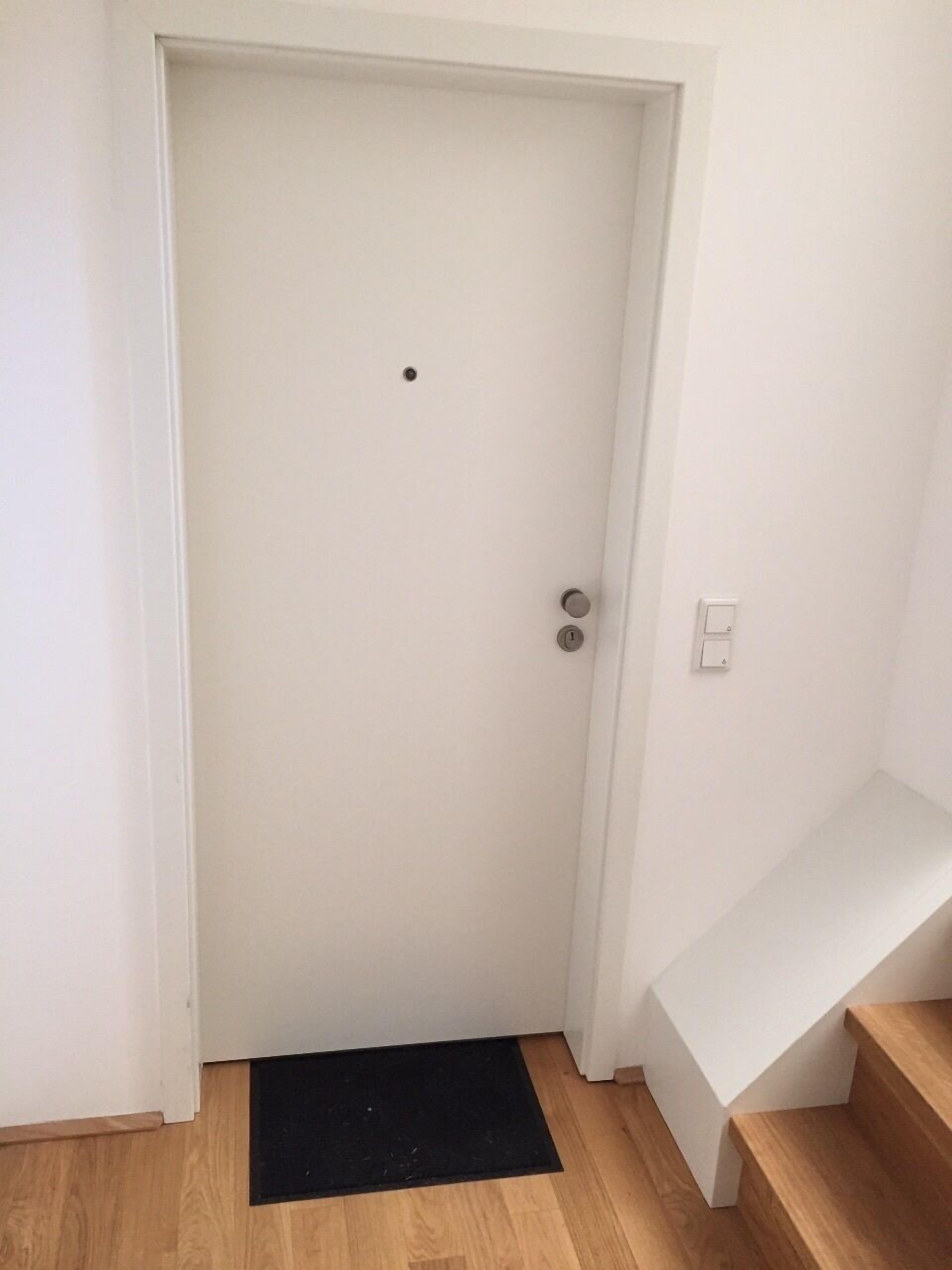 Eine weiße Tür mit Knauf und einer kleinen Öffnung darüber. Vor der Tür liegt eine schwarze Fußmatte, und rechts an der Wand befindet sich ein Lichtschalter.