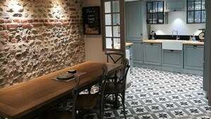 Un coin repas rustique avec une table et des chaises en bois, un mur en pierre et une cuisine dotée de meubles gris et d’un sol carrelé.
