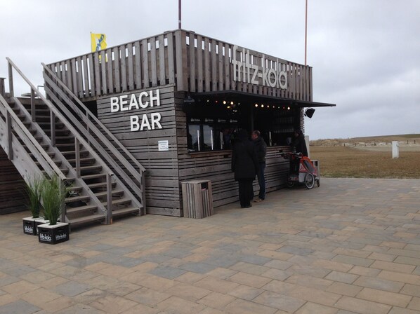 A beach bar with a wooden exterior, a staircase leading up, and a bike parked outside. - Apartment with lake views right at the Pearl Bay! (Büsum)