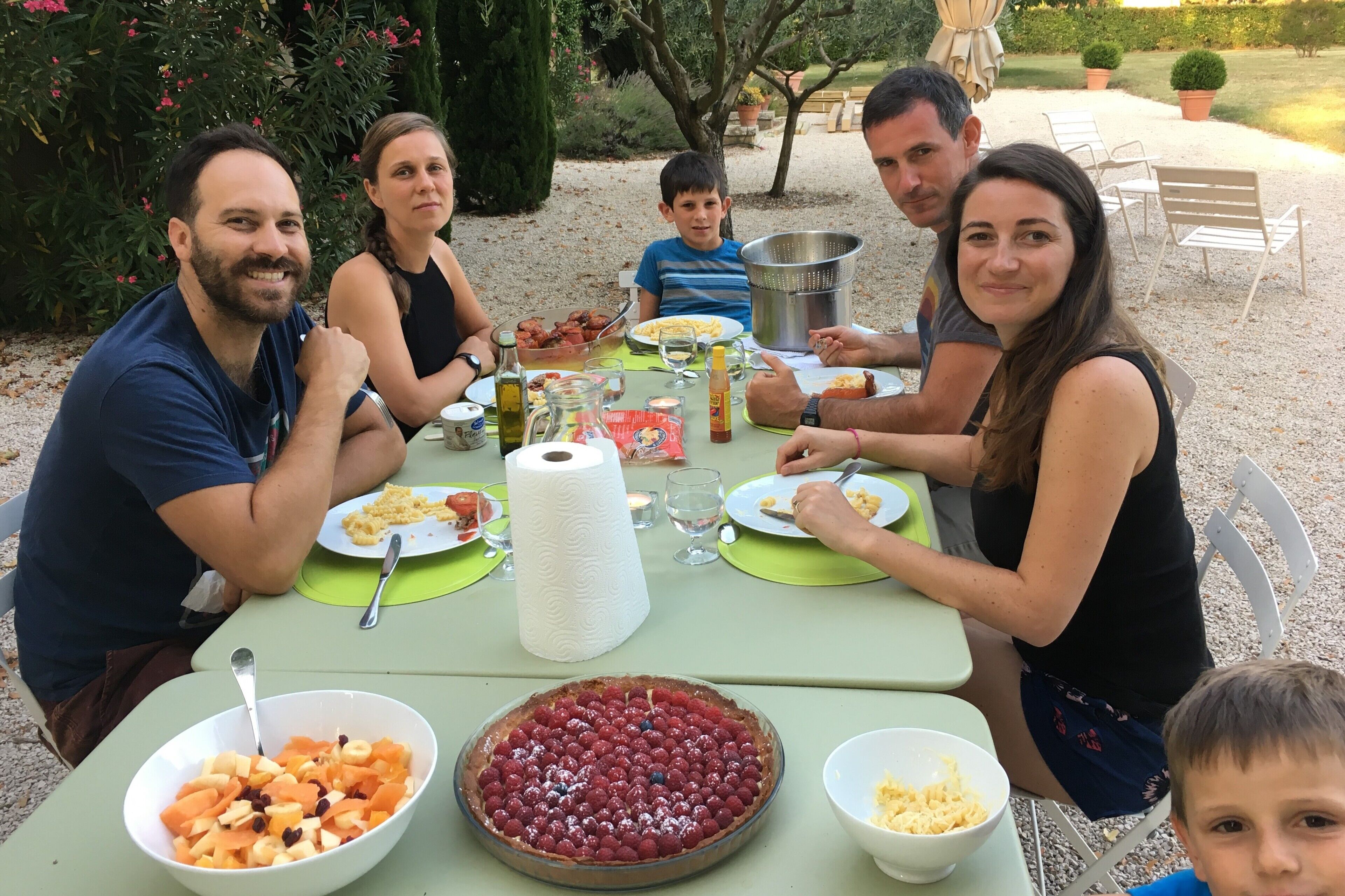 Une famille est assise à une table dressée à l’extérieur pour le repas, avec une tarte et des bols de fruits.