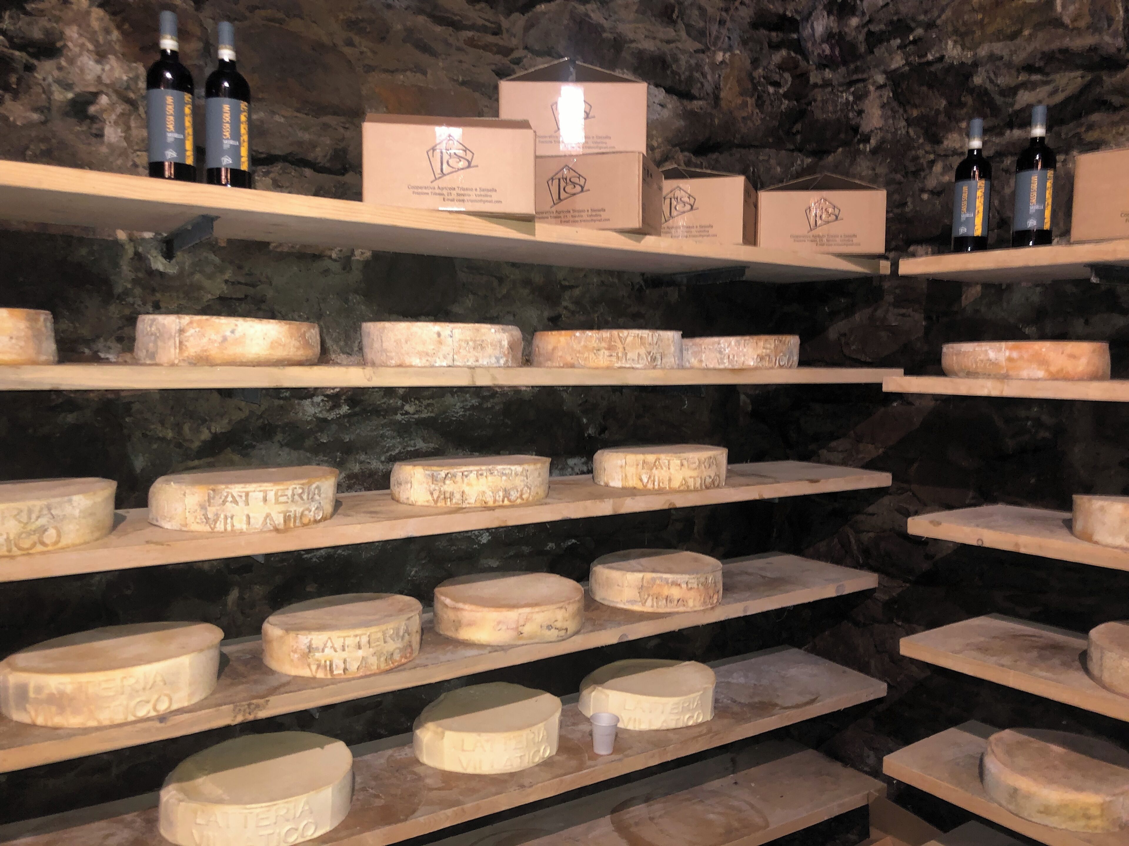 Shelves with cheese wheels and wine bottles in a cellar.