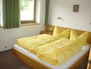 Una cama doble con colcha amarilla, cabecera de madera, mesita de noche con lámpara y ventana con cortinas verdes.