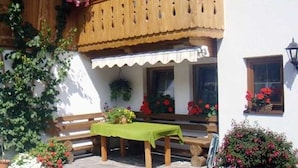 Un balcon en bois avec une table verte et des bancs, entouré de plantes et de fleurs en pot.
