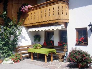 Un balcón de madera con una mesa y bancos verdes, rodeado de macetas con plantas y flores.