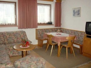 Una habitación con un sofá floral, una pequeña mesa redonda, una mesa de comedor con sillas, un televisor y una ventana con cortinas.