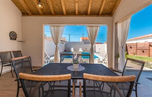 Un porche cubierto con una mesa y sillas, una piscina al fondo y vistas a una zona residencial.
