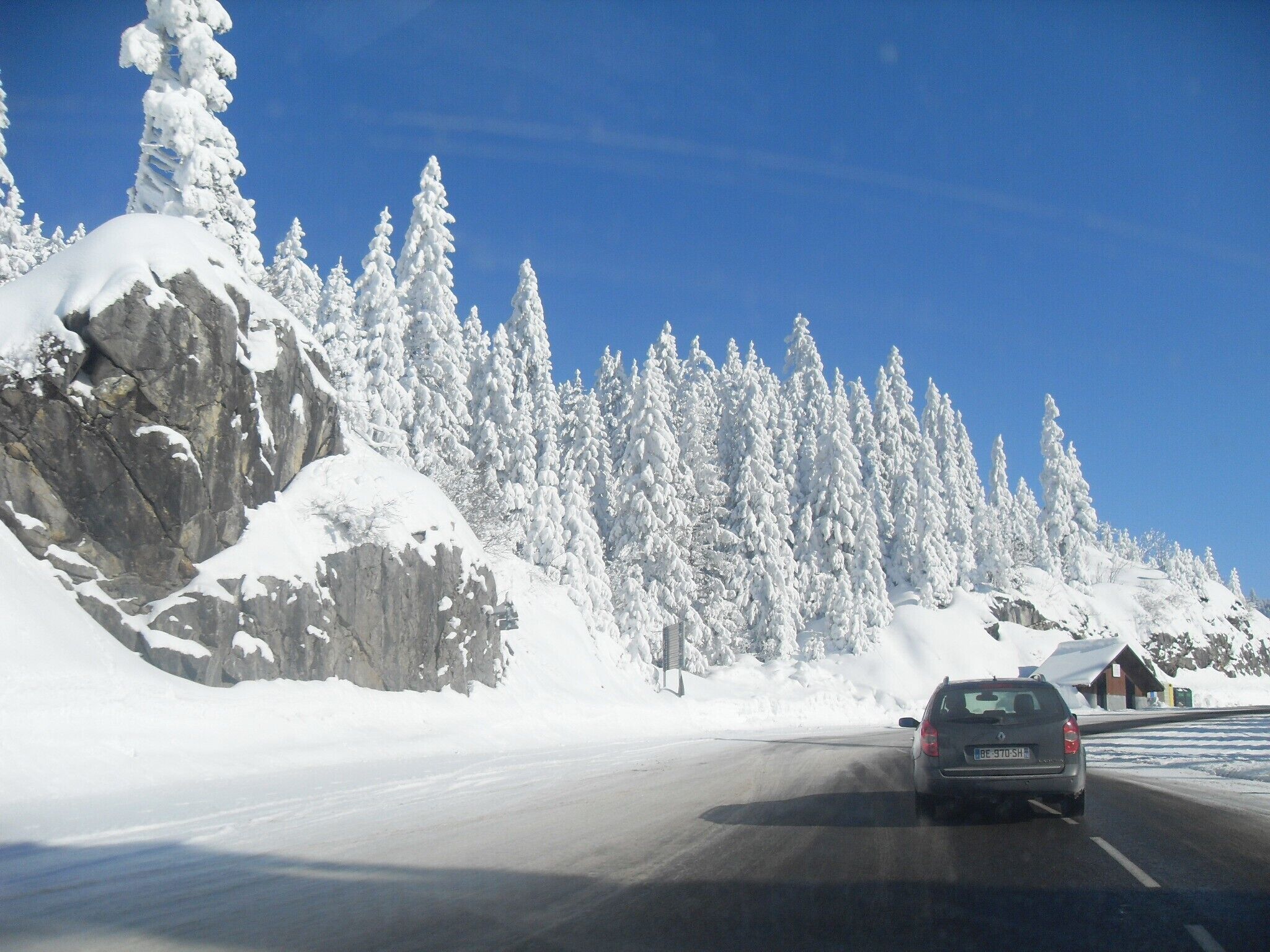 Une voiture circule sur une route enneigée, avec une chaîne de montagnes en arrière-plan.